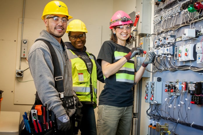 Photo of three young adults, presumably electrical trainees in safety gear working at an electrical panel.