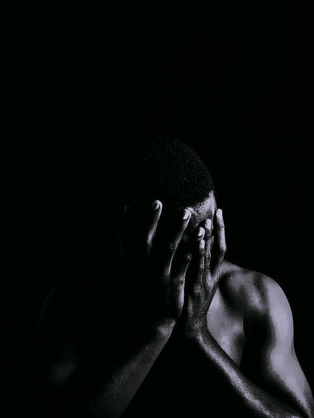 Black and white photograph of a dark skinned man with his head in his hands indicating stress.